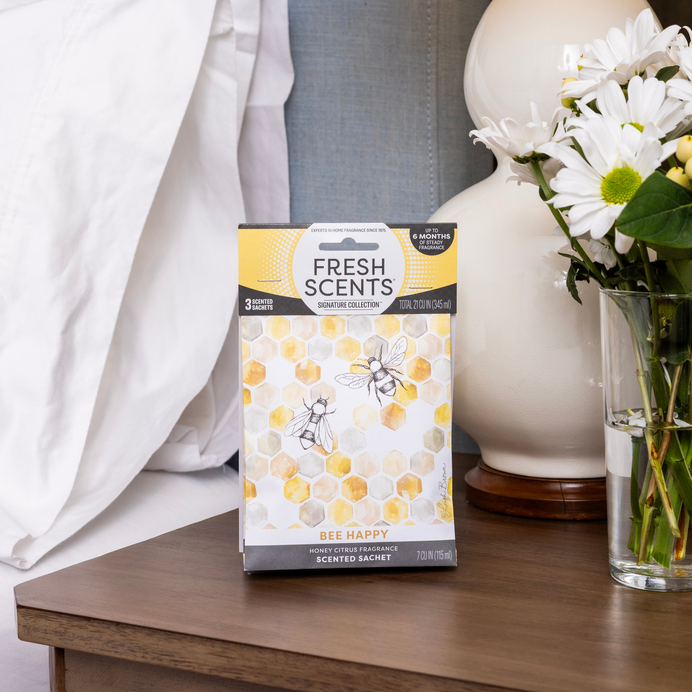 A Fresh Scents Bee Happy - Sachet 3pk with honeycomb and bee design rests on a bedside table next to a lamp and daisies, providing long-lasting, flameless fragrance as an air freshener beside the neatly made bed.