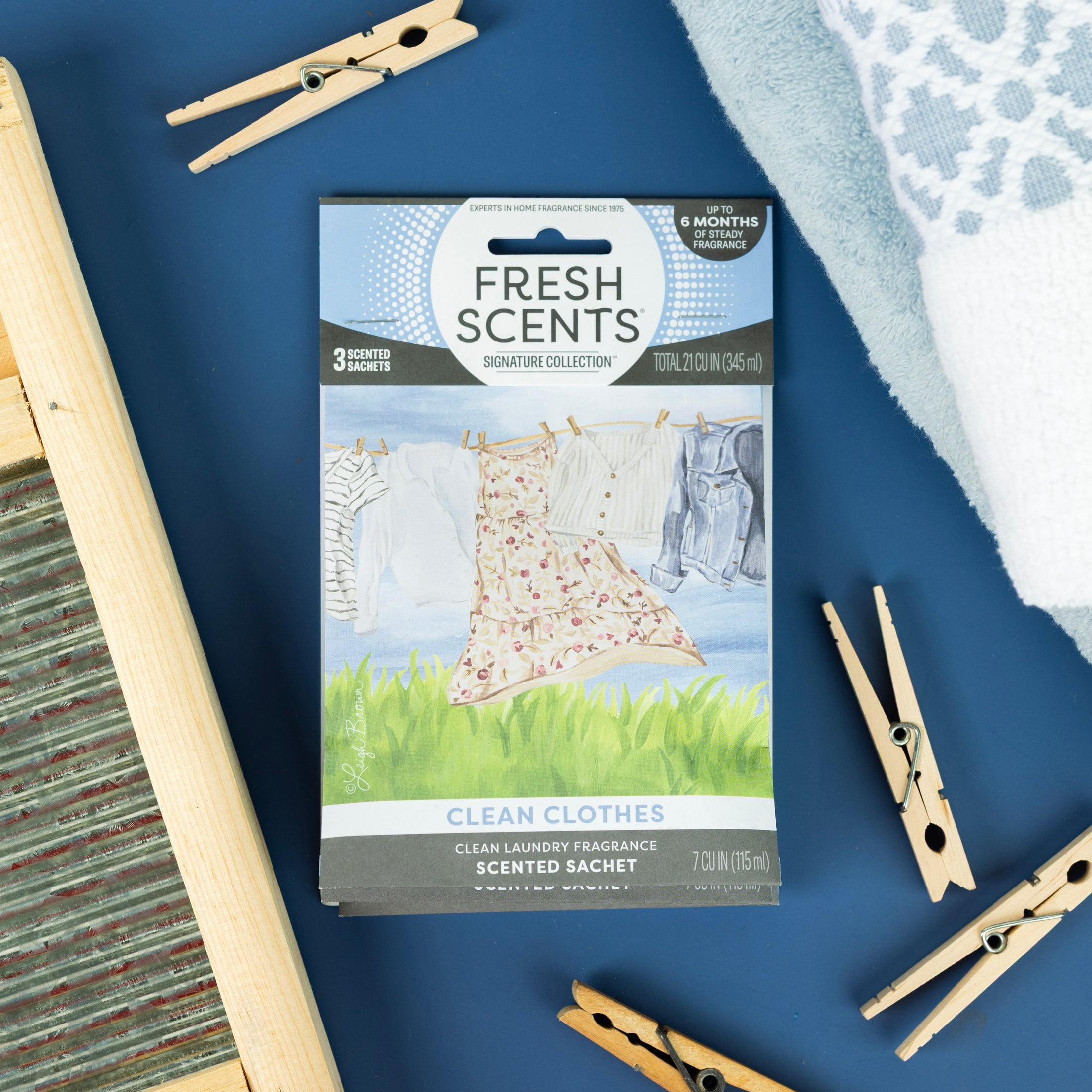 A Fresh Scents Clean Clothes - Sachet 3pk lies on a blue surface with clothespins and a washboard. This long-lasting sachet freshens your space with flameless fragrance and features a clothesline-themed illustration.