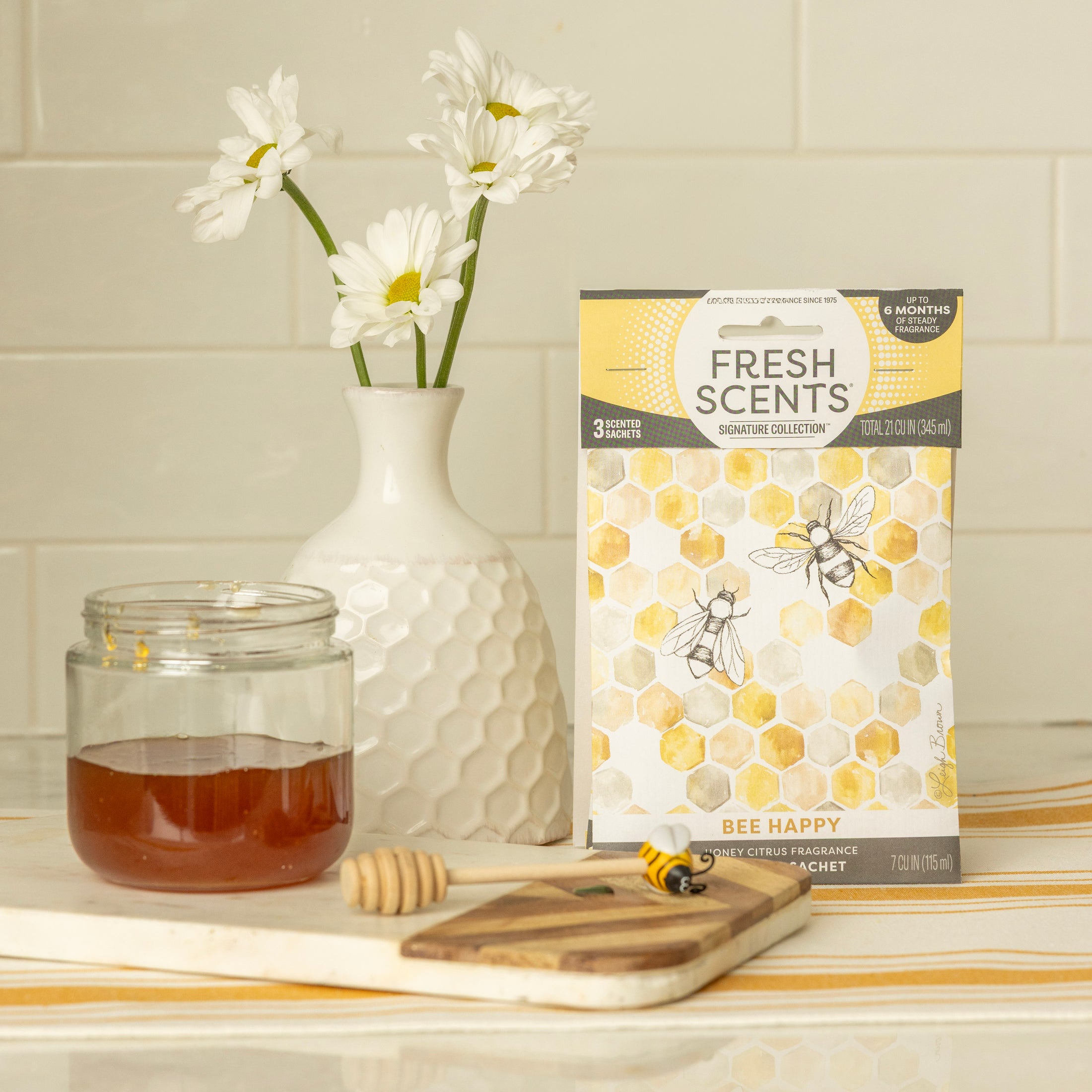 A honey jar with dipper, bee figurine, white vase with daisies, and Fresh Scents Bee Happy - Sachet 3pk featuring bee and honeycomb designs create a charming kitchen display with lasting fragrance.