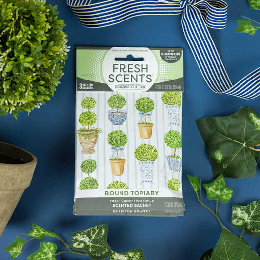 A package of Fresh Scents Round Topiary - Sachet 3pk rests on a blue surface, surrounded by potted plants, ivy leaves, and a blue-and-white striped ribbon. The packaging displays illustrations of topiary plants.