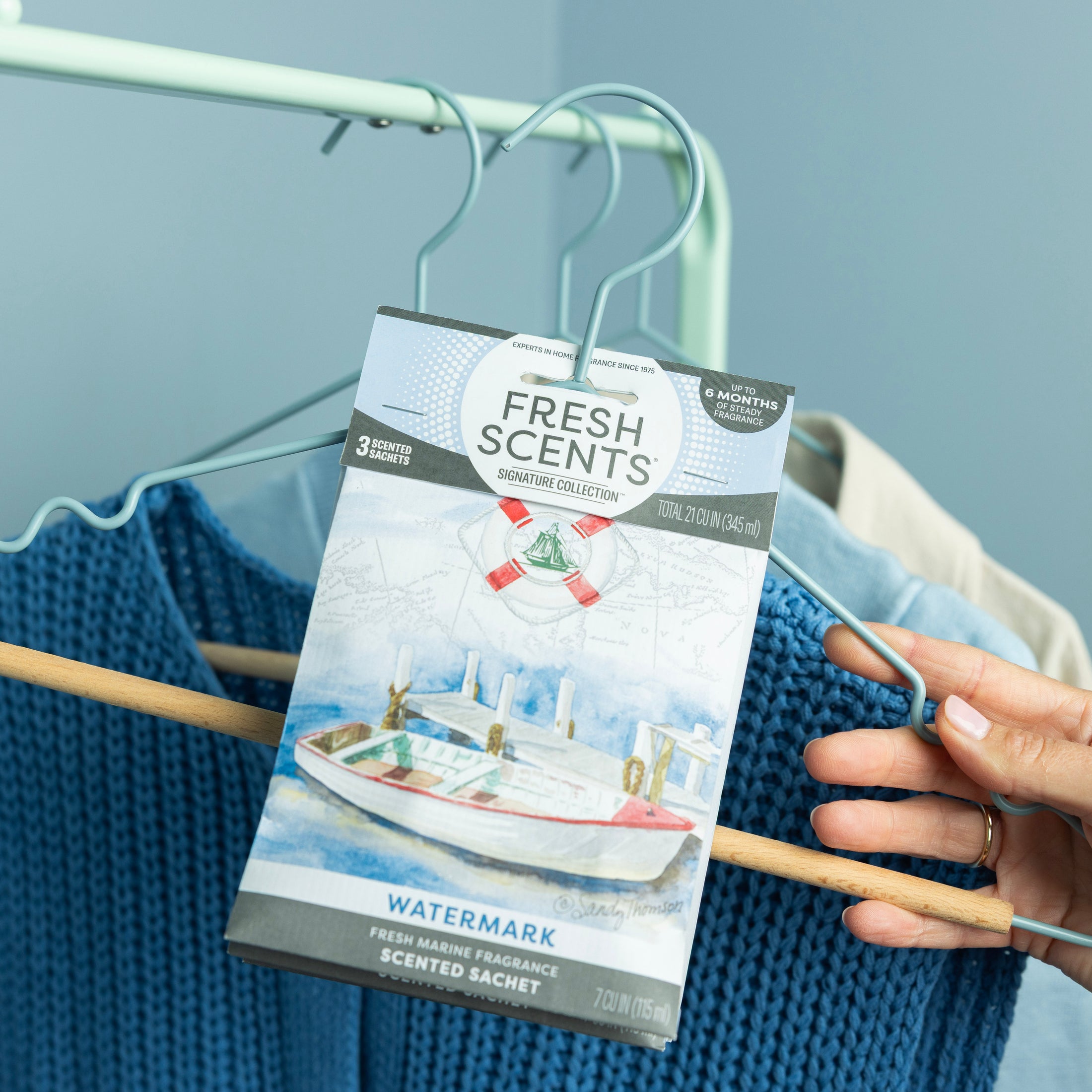 A hand hangs a Fresh Scents Watermark - Sachet 3pk, a long-lasting scented sachet, on a clothes hanger with blue and beige clothing against a light blue background.