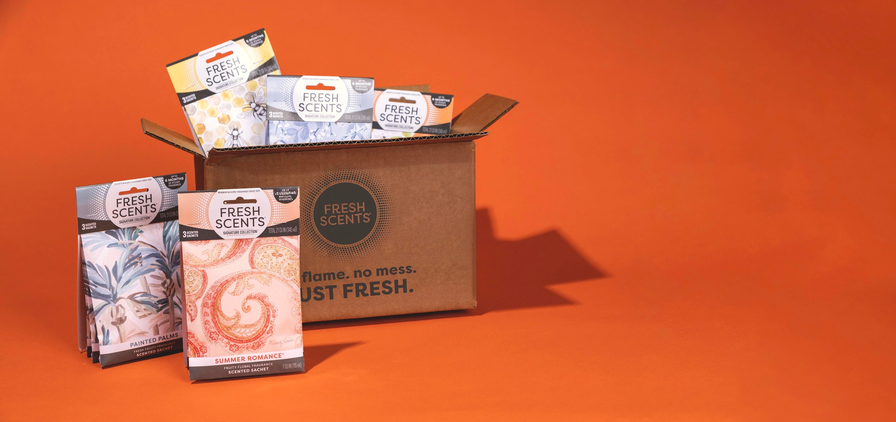 A brown box labeled Fresh Scents sits on an orange background, with various colorful Fresh Scents fragrance sachets displayed around and inside the box.