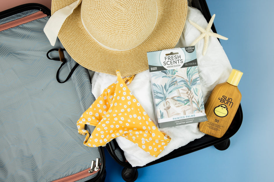 An open suitcase contains a wide-brimmed straw hat, floral yellow bikini bottoms, a starfish, sunscreen, and a scented sachet pack, all laid on a white towel against a blue background.