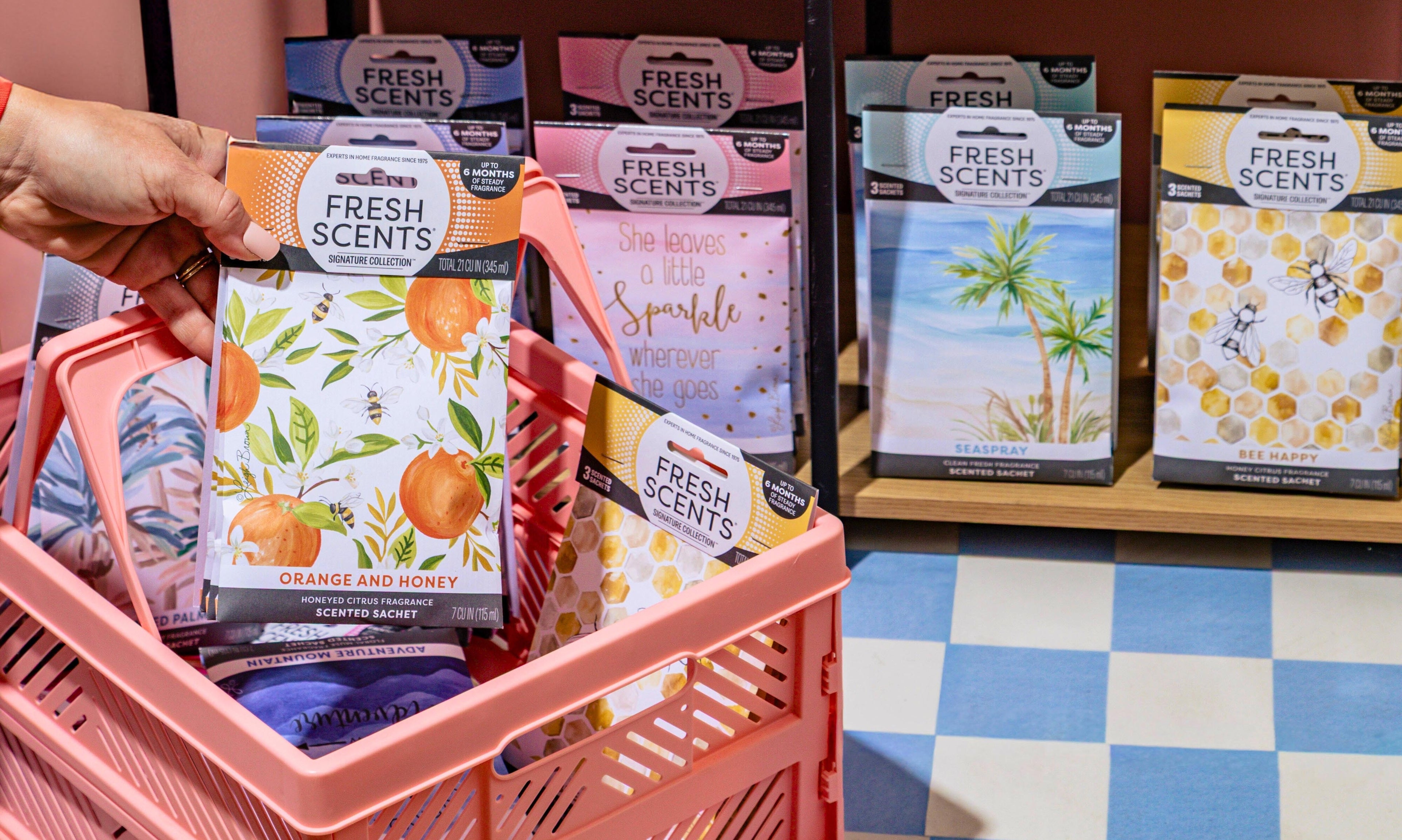 A hand places a package of Fresh Scents orange and honey scented sachets into a pink shopping basket. More sachets in various scents are displayed on shelves in the background.