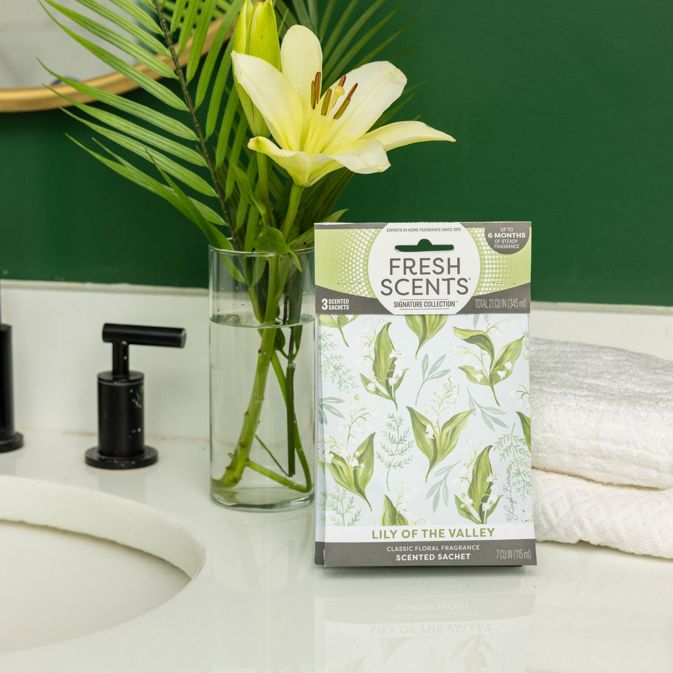 A box of Fresh Scents Lily of the Valley - Sachet 3pk, a flameless air freshener with long-lasting fragrance, sits on a bathroom counter beside yellow lilies, a black faucet, a sink, and folded white towels against a green wall.