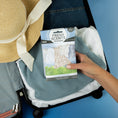 Load image into Gallery viewer, A hand places a Fresh Scents Clean Clothes - Sachet 3pk, a long-lasting air freshener, into an open suitcase with folded clothes and a straw hat with a cream-colored ribbon, set against a blue background.

