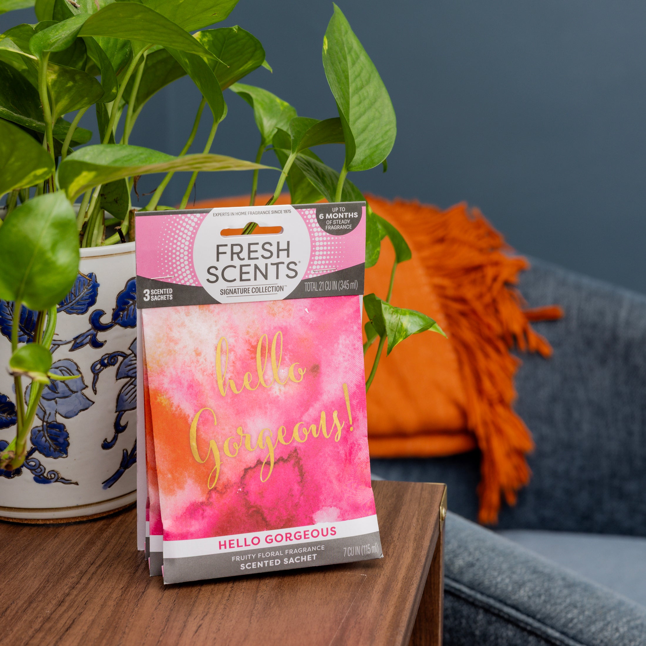 A Fresh Scents Hello Gorgeous - Sachet 3pk sits on a wooden table by a potted plant, offering long-lasting fragrance as a flameless air freshener. A blue sofa and orange pillow are visible in the background.