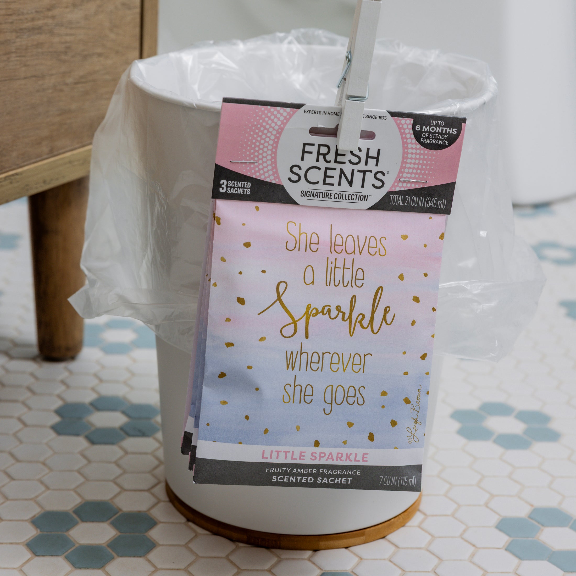 A white trash can lined with a plastic bag holds a Fresh Scents Little Sparkle - Sachet 3pk, labeled "She leaves a little sparkle wherever she goes," offering lasting fragrance in a bathroom with a tiled floor.