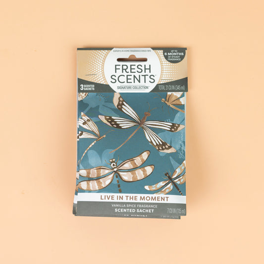 The Fresh Scents Live in the Moment - Sachet 3pk features dragonfly art on a blue background, offers long-lasting vanilla spice fragrance, and displays "LIVE IN THE MOMENT," shown on a peach-colored surface.