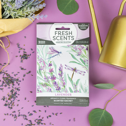 Fresh Scents Lavender - Sachet 3pk, a long-lasting air freshener in a floral box with dragonflies, is displayed on purple next to lavender flowers, scattered sachet contents, green leaves, and a gold watering can.