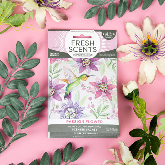 A Fresh Scents Passion Flower - Sachet 3pk rests on a pink background with faux green leaves and purple blooms. The packaging features a hummingbird and purple flowers, offering flameless fragrance.