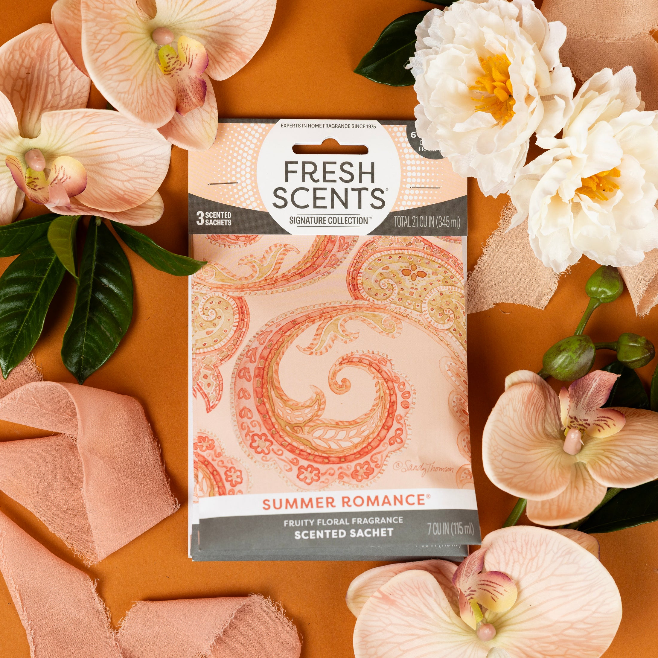 The Fresh Scents Summer Romance - Sachet 3pk rests on a brown surface among pale pink flowers, white roses, green leaves, and a soft pink ribbon—a flameless, long-lasting air freshener for any space.
