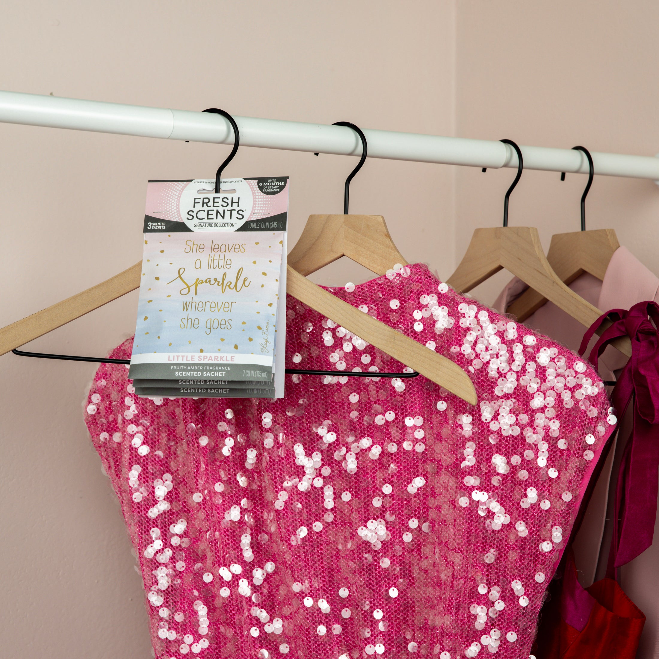 A bright pink sequin dress hangs on a rack with a Fresh Scents Little Sparkle - Sachet 3pk, adding long-lasting fragrance with the message "She leaves a little sparkle wherever she goes." More pink dresses hang in the background.
