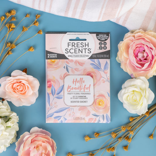 A Fresh Scents "Hello Beautiful - Sachet 2pk" sits on a blue surface among pastel artificial flowers and dried stems, offering a charming and fragrant way to freshen any room.