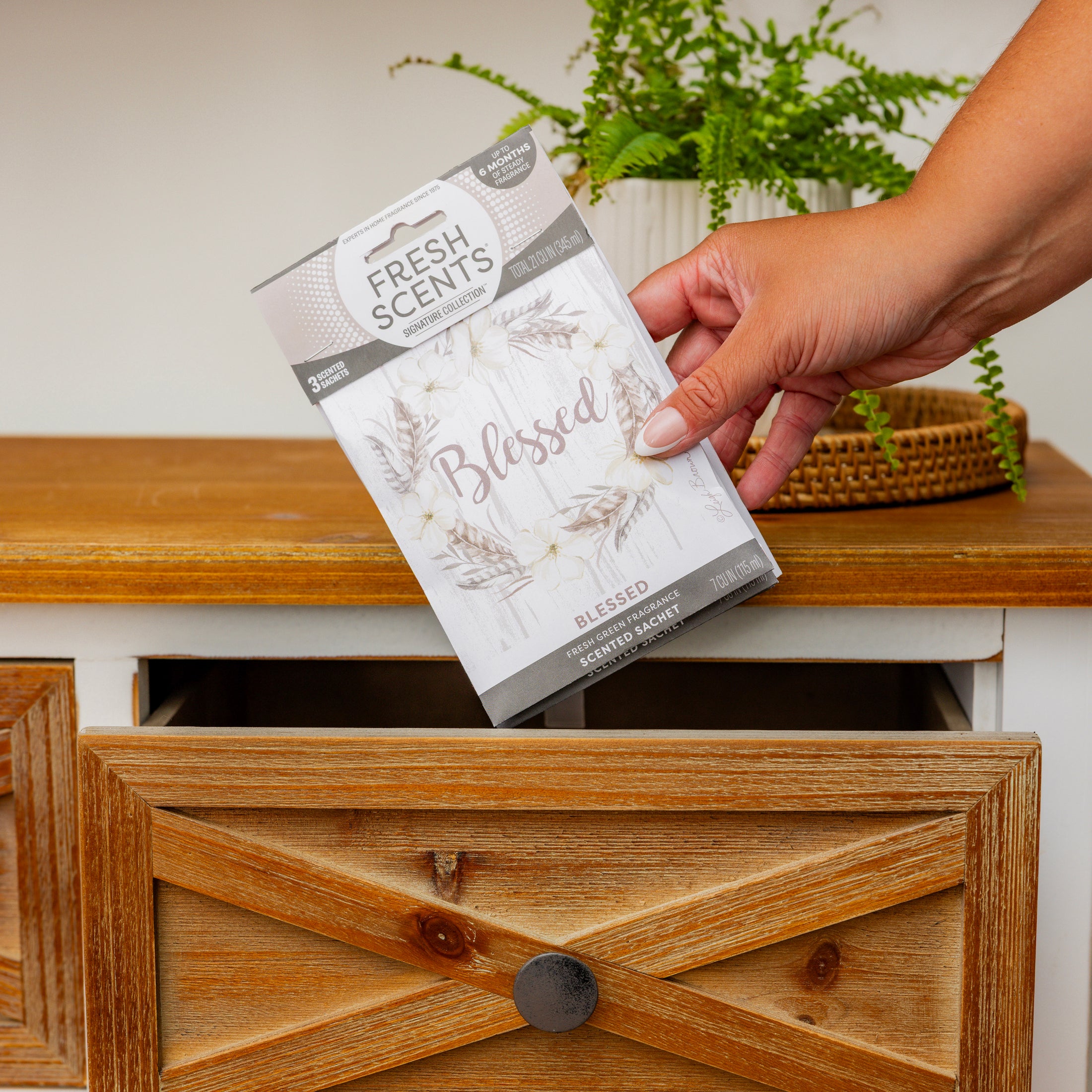 A hand places a Fresh Scents Blessed Wreath 3pk scented sachet into an open wooden drawer. A green potted plant sits nearby, offering a flameless, long-lasting air freshener for your space.