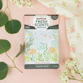 Load image into Gallery viewer, A package of Fresh Scents Baby Powder - Sachet 3pk, a long-lasting air freshener, lies on a pink surface with green leaves and baby's breath flowers. The packaging features pastel flower illustrations and floral-patterned cloth accents.