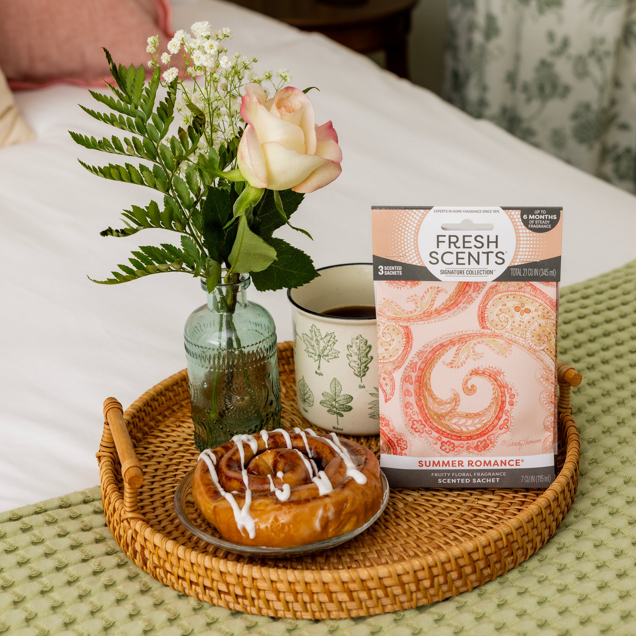 A wicker tray on a bed holds a cinnamon roll, coffee mug, small vase with a rose and greenery, and a Fresh Scents Summer Romance Sachet 3pk—a flameless fragrance for subtle, long-lasting freshness.