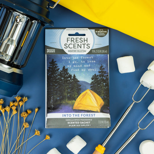 A flat lay of camping items including a lantern, marshmallows, roasting sticks, dried flowers, and a Fresh Scents Into the Forest - Sachet 3pk featuring a yellow tent illustration under a night sky.