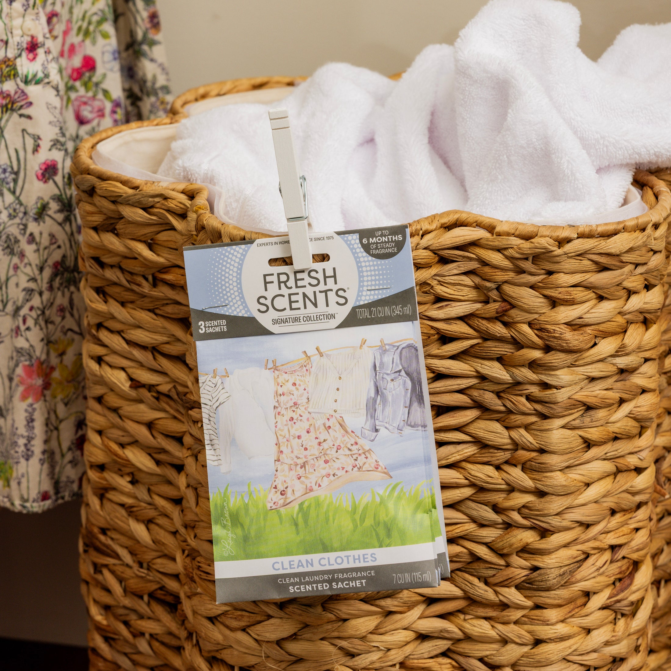 A woven laundry basket filled with white towels has a Fresh Scents Clean Clothes - Sachet 3pk clipped to the front for flameless fragrance. A floral dress hangs nearby.