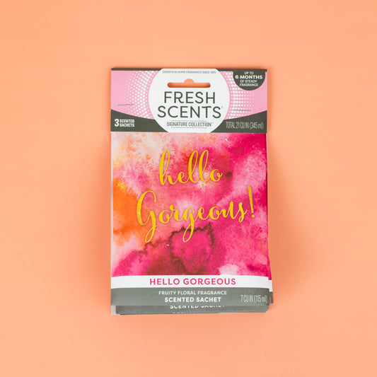 The Fresh Scents Hello Gorgeous - Sachet 3pk features pink and orange packaging with gold script on a peach background. This flameless air freshener delivers a long-lasting fruity floral scent and includes three scented sachets.