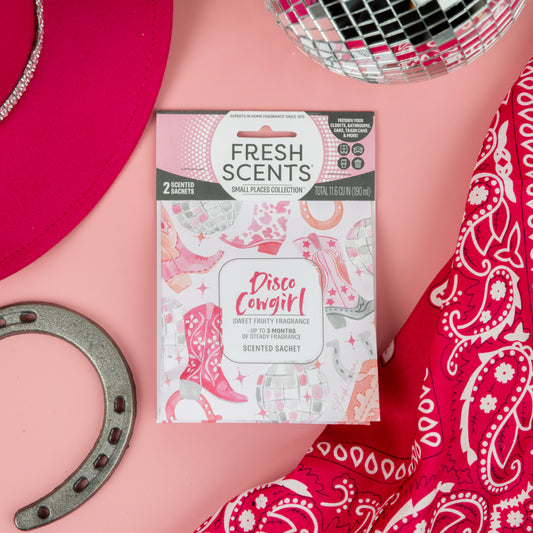 Fresh Scents Disco Cowgirl - Sachet 2pk sits on a pink background, surrounded by a red cowboy hat, silver horseshoe, disco ball, and red bandana—the perfect flameless air freshener for any small space!.