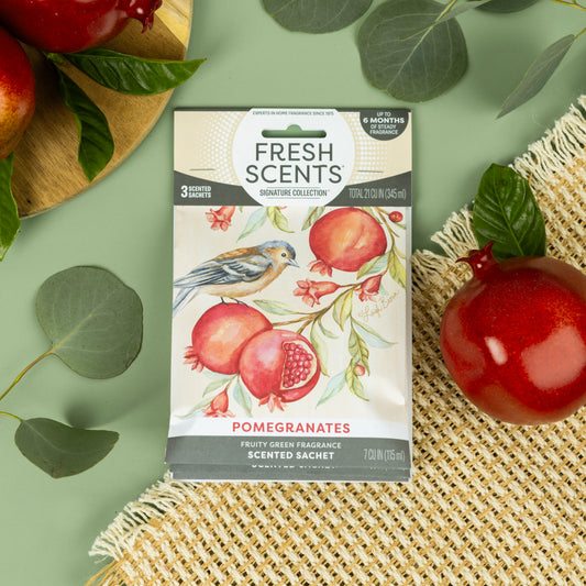 A Fresh Scents Pomegranate - Sachet 3pk with pomegranate art and a bird rests on a woven mat. Real pomegranates, green leaves, and a gold-rimmed plate complete the display, bringing long-lasting fragrance to the scene.