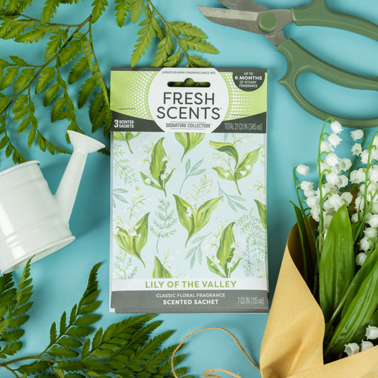 A Fresh Scents Lily of the Valley - Sachet 3pk, a flameless air freshener with long-lasting fragrance, sits on a blue surface among fern leaves, garden shears, a white watering can, and lily of the valley flowers wrapped in brown paper.