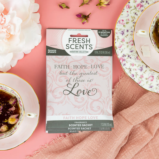 The Faith Hope Love - Sachet 3pk by Fresh Scents rests on a pink surface with teacups, floral tea, a pink flower, and napkin—ideal for long-lasting fragrance or as a flameless air freshener.
