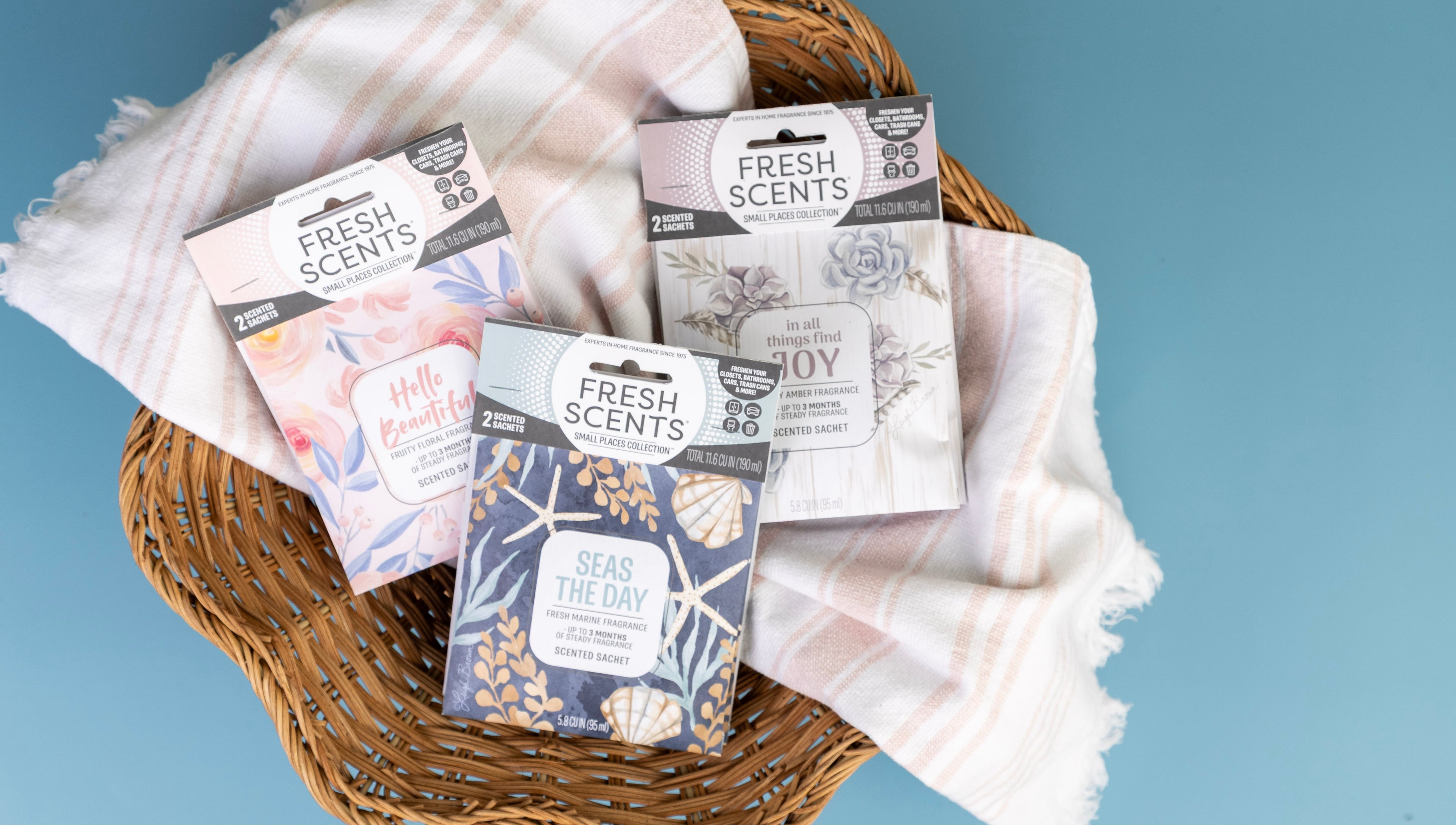 Three Fresh Scents sachets with floral designs in a wicker basket lined with a pink and white striped cloth, set against a blue background.
