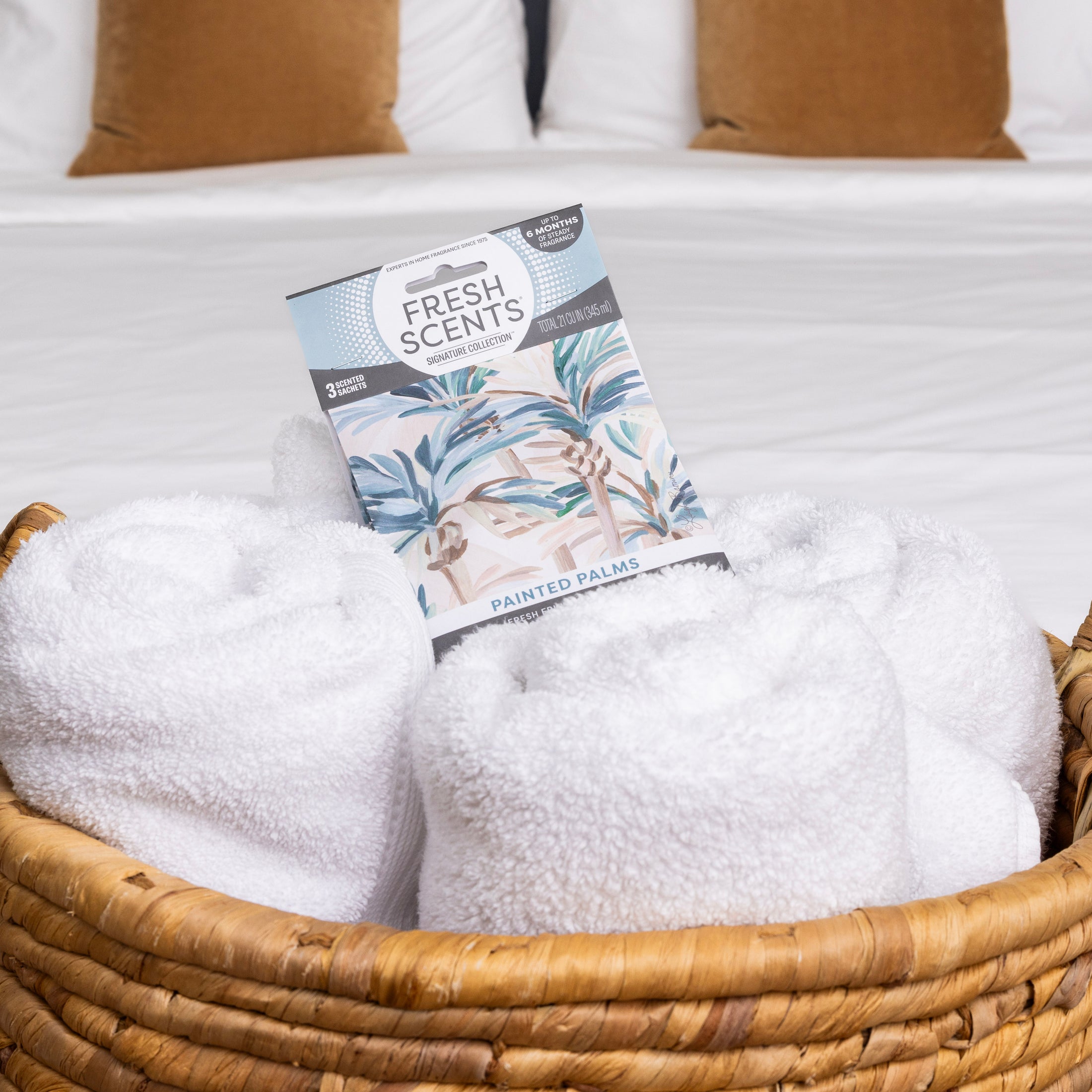 A woven basket with rolled white towels and a Fresh Scents Painted Palm - Sachet 3pk, a flameless fragrance option, rests on a white bed with two brown pillows in the background.