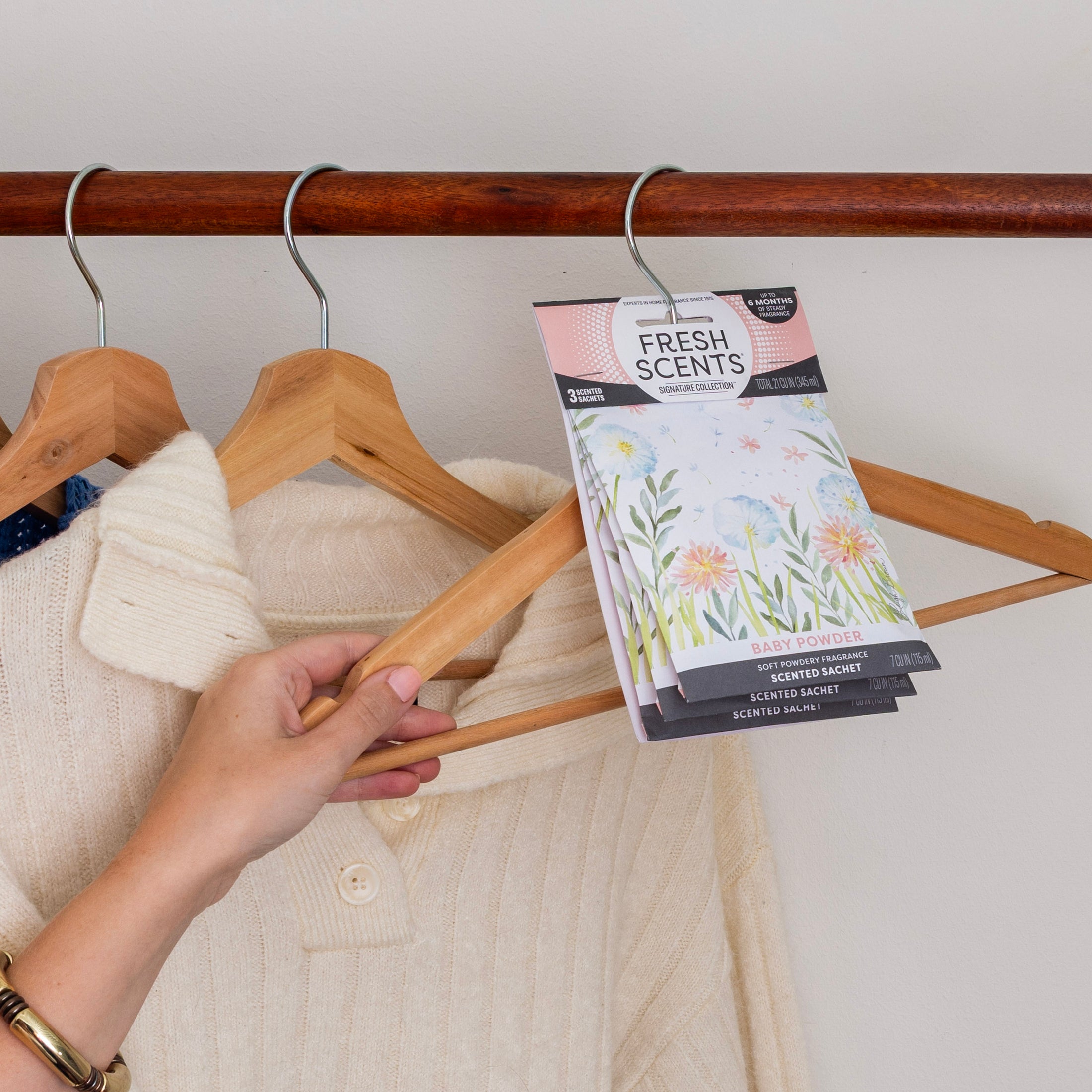 A hand hangs a cream knit sweater and a Fresh Scents Baby Powder - Sachet 3pk, a flameless fragrance, from a wooden hanger on a closet rod.