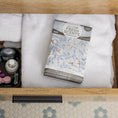 Load image into Gallery viewer, An open wooden drawer holds two folded white towels, a Fresh Scents White Cotton - Sachet 3pk for long-lasting fragrance, and a small organizer with nail polish and grooming items, above a hexagon-tiled floor.
