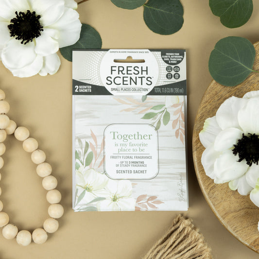 A Fresh Scents Together - Sachet 2pk with floral designs rests on a beige surface amid white flowers, green leaves, a wooden beaded bracelet, and a fringed coaster—ideal fragrance sachets for small spaces as flameless air fresheners.