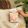 Load image into Gallery viewer, A hand places a Fresh Scents Summer Romance - Sachet 3pk, featuring a pink paisley design, on a beige pillow to provide flameless fragrance near a green plant and woven basket chair.
