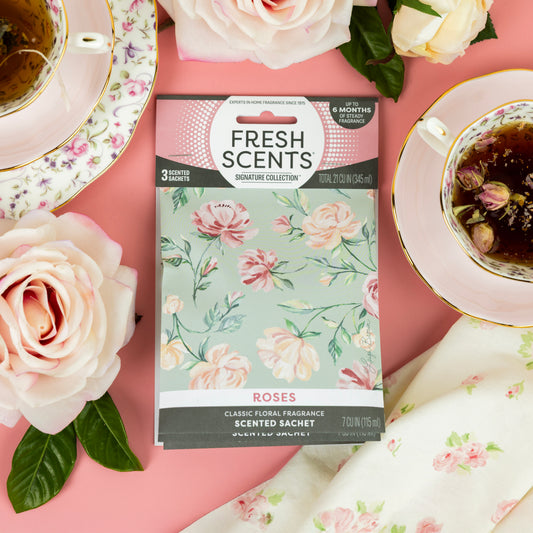 Fresh Scents Roses - Sachet 3pk rests on a pink surface, surrounded by floral teacups, pink roses, and a printed white napkin—an elegant way to enjoy flameless fragrance and long-lasting freshness.