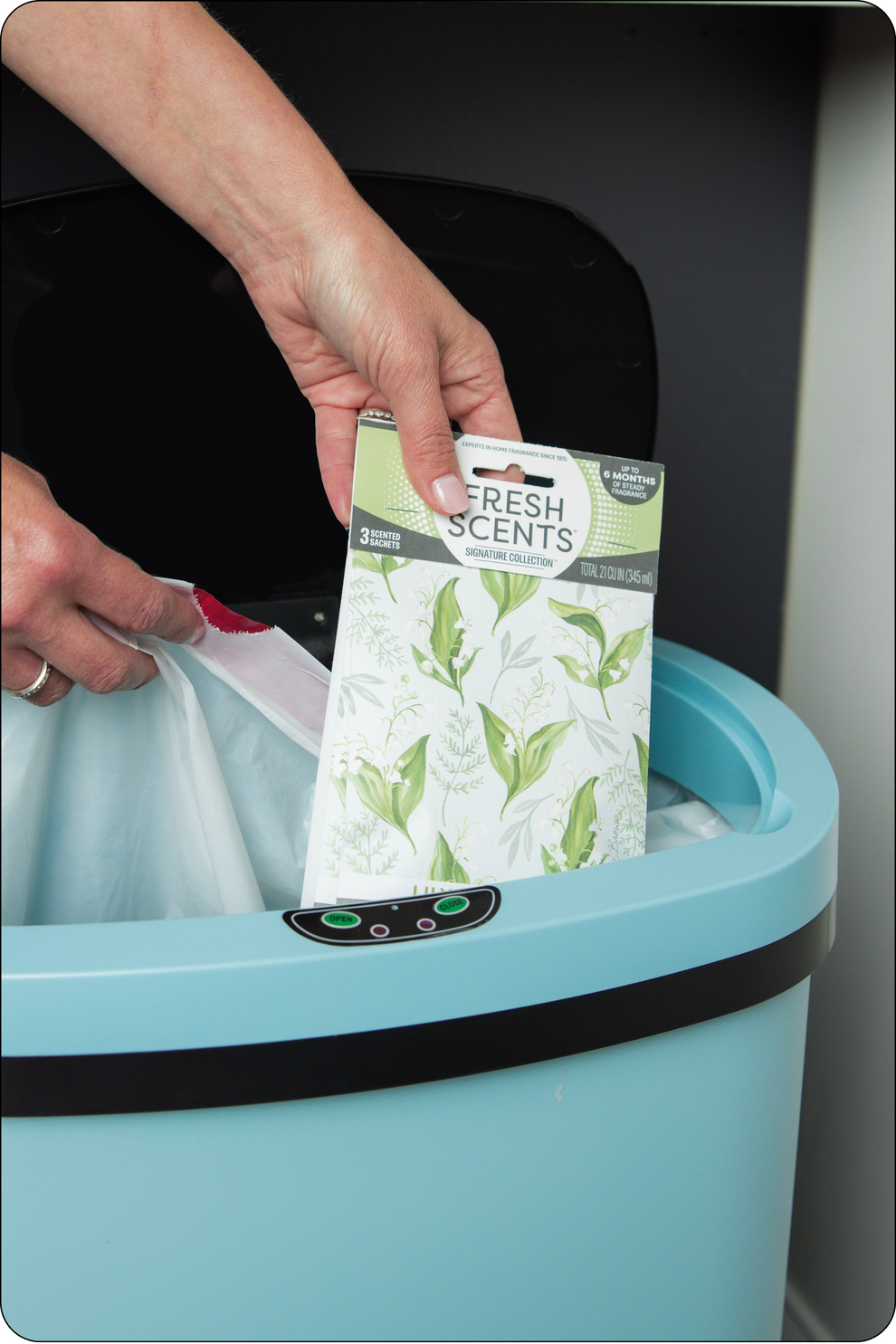 A person places a Fresh Scents sachet into a blue trash can lined with a white garbage bag. The trash can lid is open, revealing the inside.