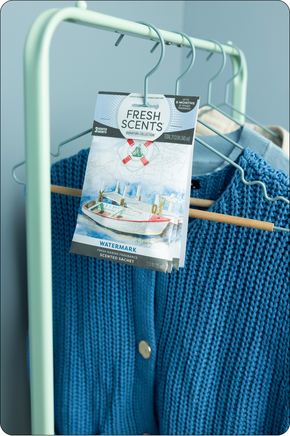 A blue knit sweater hangs on a light green clothing rack, with a Fresh Scents scented sachet labeled Watermark clipped to the rack in front of the sweater. Other clothing items are visible in the background.